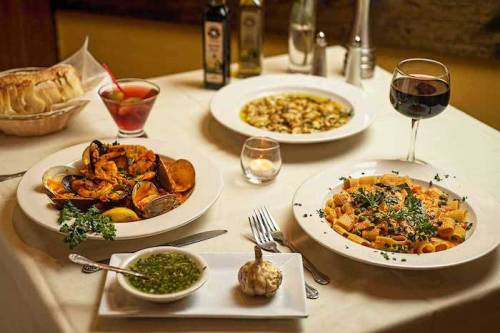 A table set with three dishes: seafood, pasta, and a green sauce, accompanied by wine and a cocktail.