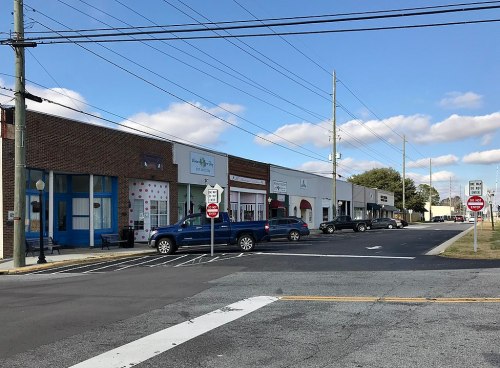 A street view of a small town with shops, parked cars, and power lines under a partly cloudy sky.