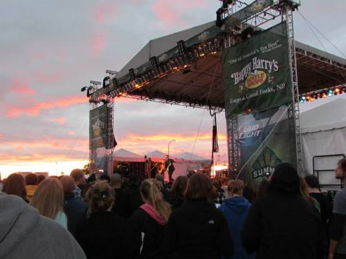 A crowd watches a live performance on stage at sunset, with colorful skies and festival banners in the background.