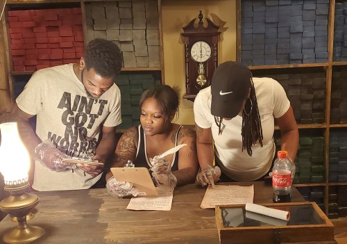 Three people in a cozy room are reviewing notes and writing on clipboards, with a vintage clock in the background.