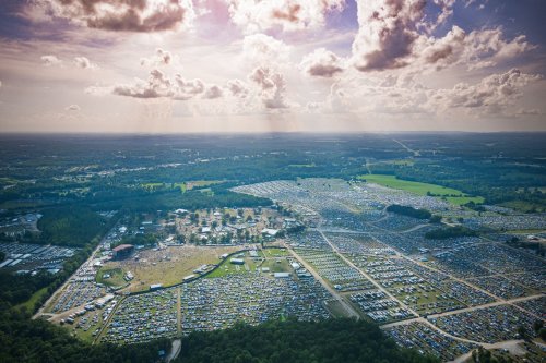 Aerial view of a large outdoor event with numerous cars parked in a vast field under a cloudy sky.