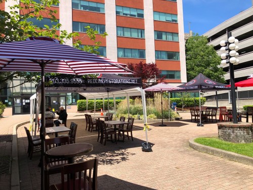 Outdoor seating area with striped umbrellas and tables, surrounded by greenery and buildings.