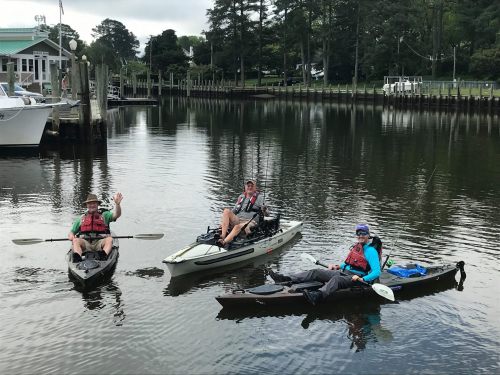 Three people in kayaks on a calm river, smiling and enjoying a sunny day, surrounded by trees and boats.