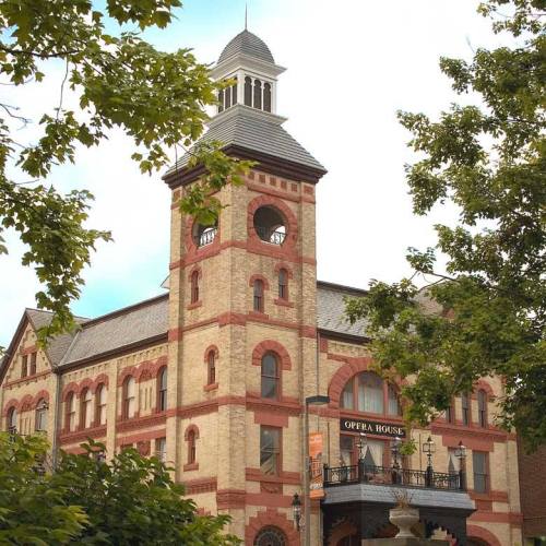 Historic building with a clock tower, featuring red brick accents and a sign reading "Opera House," surrounded by trees.