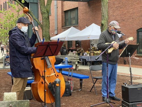 Two musicians perform outdoors: one plays a double bass, while the other strums a ukulele, both wearing masks.