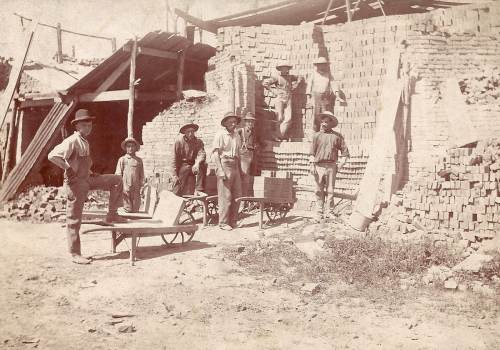 A group of seven men and a boy pose near a brick wall and wheelbarrows at a construction site.