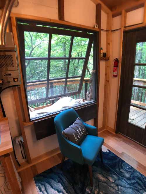 Cozy interior of a cabin with a blue chair, a window overlooking trees, and a decorative pillow that says "hello."