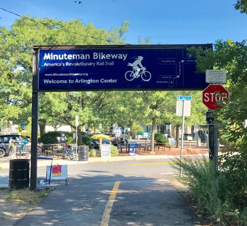 Sign for the Minuteman Bikeway welcoming visitors to Arlington Center, with trees and a stop sign in the background.