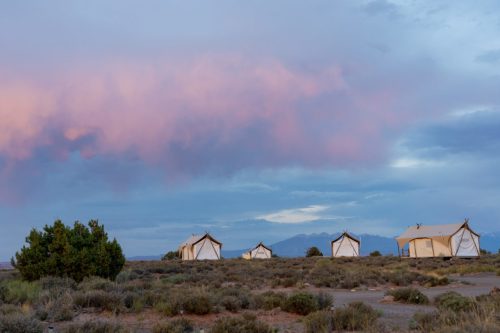 A serene landscape featuring glamping tents under a colorful sky with soft clouds and distant mountains.