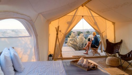 A cozy tent interior with a bed and a person sitting outside, enjoying a scenic view at sunset.
