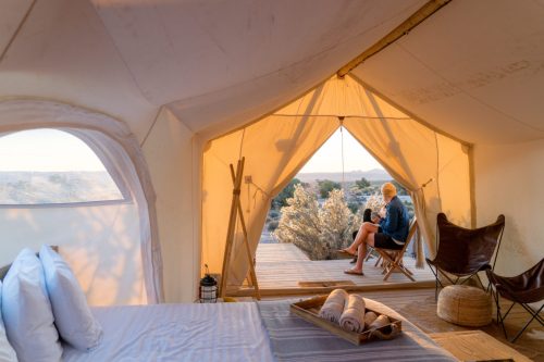 A cozy tent interior with a bed, overlooking a person sitting outside on a deck, enjoying the view at sunset.