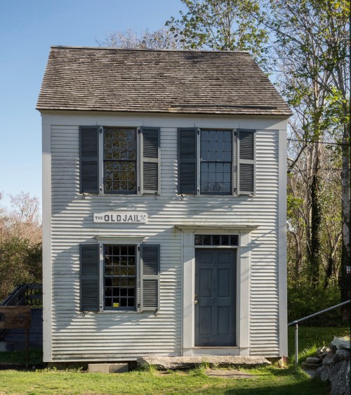 A historic two-story building with gray siding, black shutters, and a sign reading "The Old Jail." Surrounded by greenery.