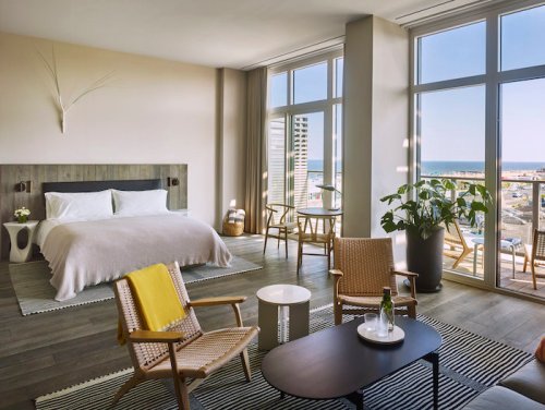 Bright hotel room with a large bed, seating area, and ocean view through floor-to-ceiling windows. Modern decor.