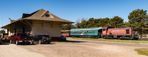 A vintage train and green passenger car beside a historic train station, with parked cars in the foreground.