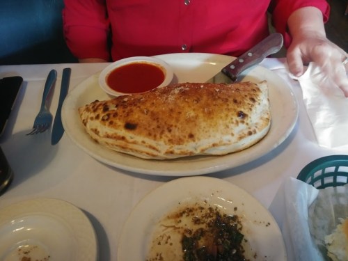 A large, golden-brown calzone on a plate, accompanied by a small bowl of red sauce and a side of chopped herbs.