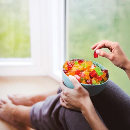 A person sitting by a window holds a bowl of colorful gummy candies, with rain visible on the glass.