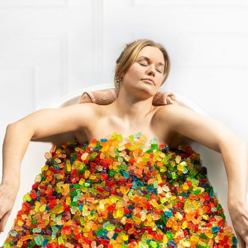 A woman relaxes in a bathtub filled with colorful gummy candies, eyes closed and a serene expression.