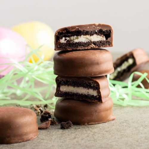 Three chocolate-covered cookies stacked, with one cut in half to reveal a creamy filling, surrounded by pastel decorations.