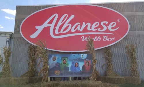A large Albanese sign with a festive display featuring people in colorful costumes and hay bales in front.