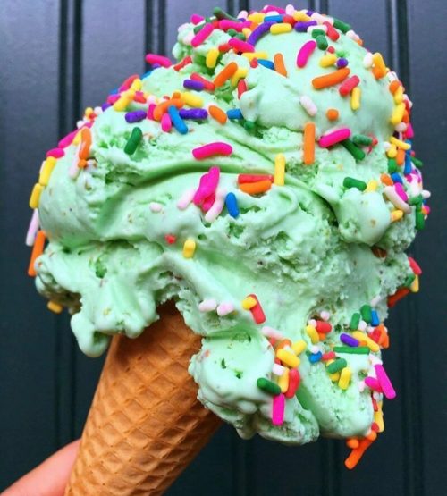 A close-up of a mint green ice cream cone topped with colorful sprinkles against a dark green background.