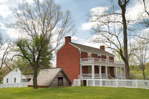 Historic red brick house with white porch, surrounded by green grass and trees, featuring a nearby wooden structure.