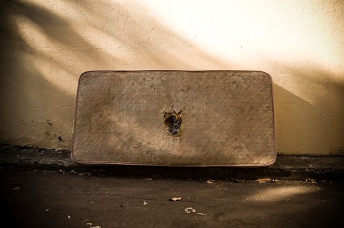 A worn-out mattress leaning against a wall, showing a large tear in the center. Sunlight casts shadows on the ground.