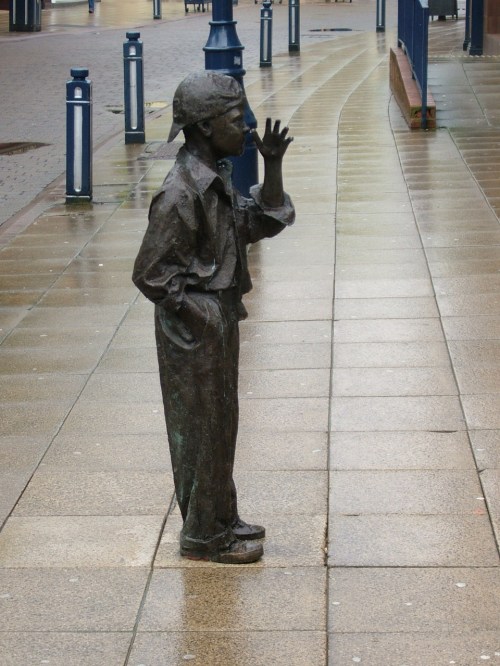 A bronze statue of a young boy in a cap, standing on a wet sidewalk, with one hand raised in a wave.
