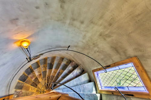 A spiral staircase with stone steps, leading up to a window with a grid pattern and a warm light fixture above.