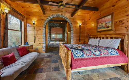Cozy wooden cabin interior featuring a log bed, quilt, stone accents, and a sofa in a warm, inviting atmosphere.
