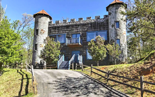 A stone castle-like house with turrets, surrounded by trees and a gravel driveway leading up to it.