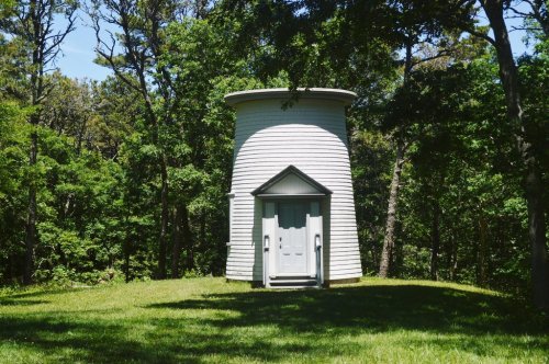 A white, cylindrical building surrounded by trees, set on a grassy area under a clear blue sky.