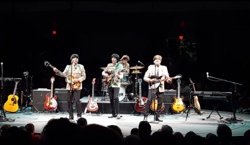A tribute band performs on stage with guitars, drums, and amplifiers, entertaining a seated audience.