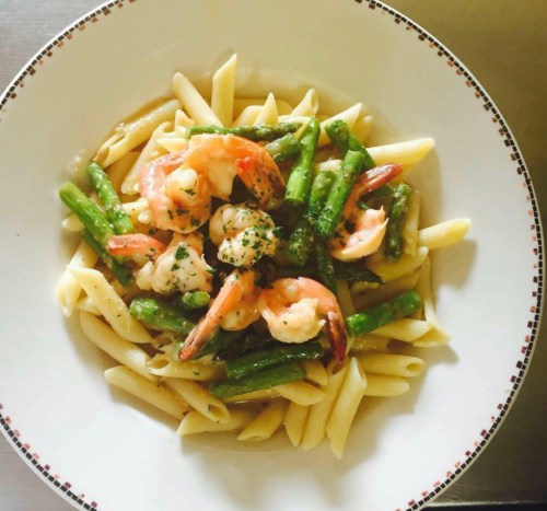 A bowl of penne pasta topped with shrimp and green asparagus, garnished with herbs.