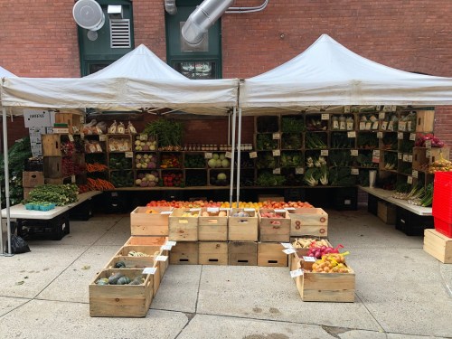 A vibrant outdoor market stall with various fresh vegetables and fruits displayed in wooden crates under white tents.
