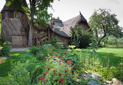 A whimsical, rustic house with a thatched roof, surrounded by vibrant flowers and lush greenery.