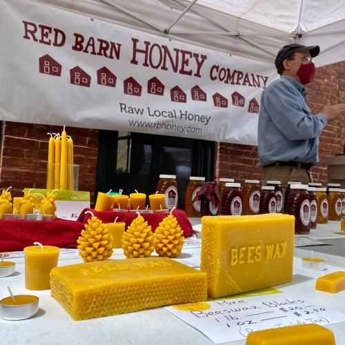 A vendor at a market displays beeswax products and honey jars under a tent for Red Barn Honey Company.