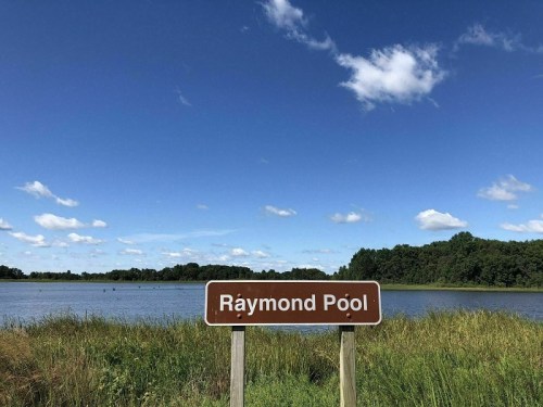 A sign reading "Ráymond Póol" in front of a calm lake surrounded by green grass and trees under a blue sky.