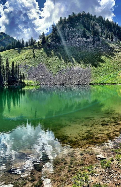A serene lake reflects a green hillside and blue sky, surrounded by trees and mountains under fluffy clouds.