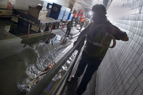 Workers in safety gear manage equipment in a dimly lit tunnel with water on the floor.