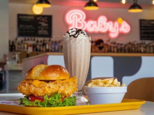 A crispy chicken sandwich with cheese, a side of macaroni, and a chocolate milkshake in a vibrant diner setting.