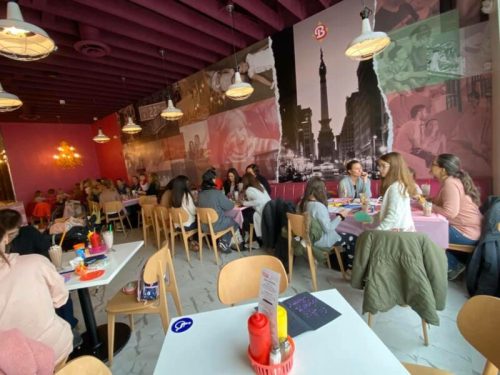 A lively restaurant interior with pink walls, tables filled with people, and decorative lighting.