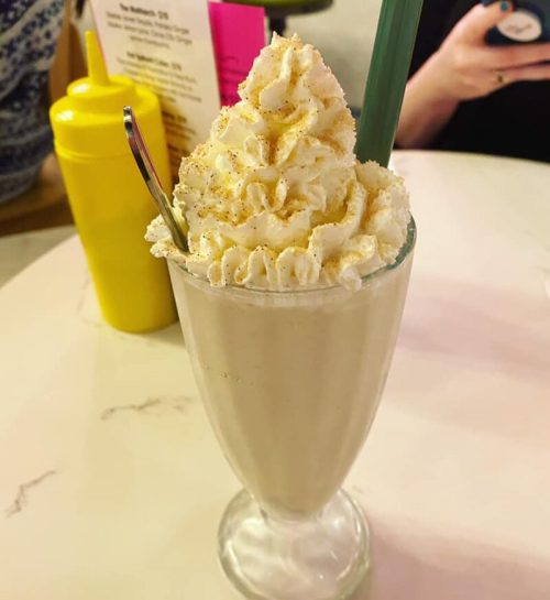 A tall glass of milkshake topped with whipped cream and a sprinkle of cinnamon, served with a spoon and straw.