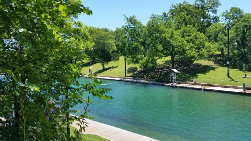 A serene canal lined with trees, featuring clear water and people enjoying the sunny day on the grassy banks.