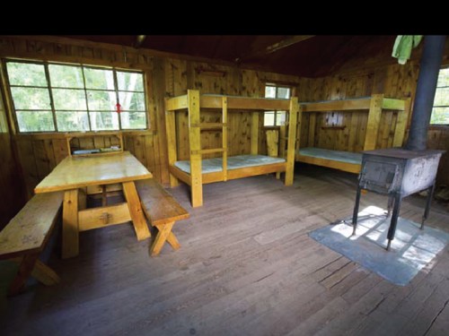Interior of a rustic cabin featuring wooden bunk beds, a table, and a stove, with large windows letting in natural light.