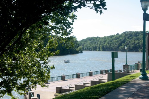 A serene river scene with a small boat, surrounded by lush greenery and a paved walkway along the water's edge.