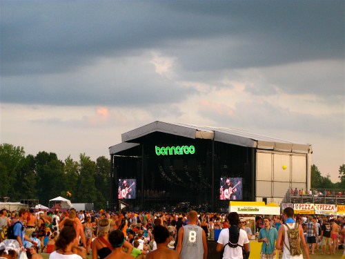 A large crowd gathers at a music festival stage under a cloudy sky, with food vendors visible in the foreground.
