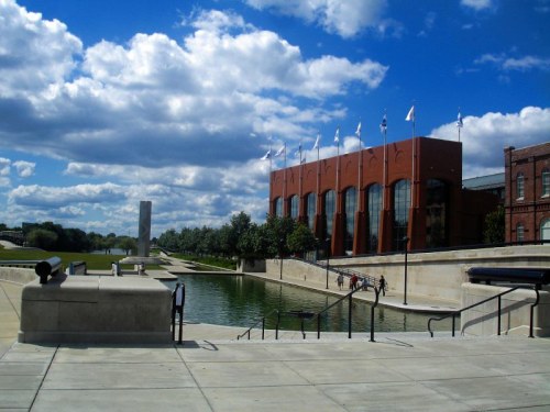 A modern building with flags beside a calm waterway, under a blue sky with fluffy clouds. Steps lead down to the water.
