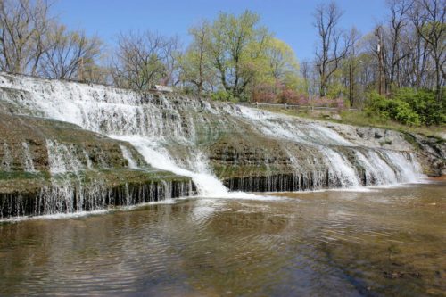 A serene waterfall cascading over rocky ledges, surrounded by lush greenery and clear blue skies.