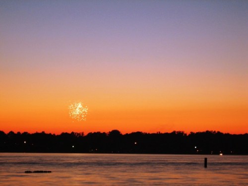 Fireworks sparkle in the sky above a calm lake at sunset, with silhouettes of trees and distant lights along the shore.