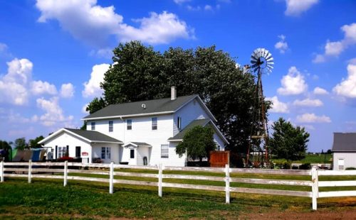 A white farmhouse with a windmill nearby, surrounded by green trees and a white fence under a blue sky with clouds.